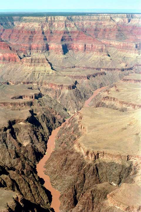 Aerial view looking up a muddy Colorado River with dark brown cliffs rising to a flat plateau, then to sheer reddish cliffs in the distance.