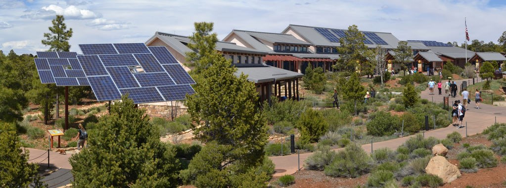 people walking on a sidewalk that winds its way around plaza vegetation towards a group of modern buildings. On the left, two large solar panel arrays.