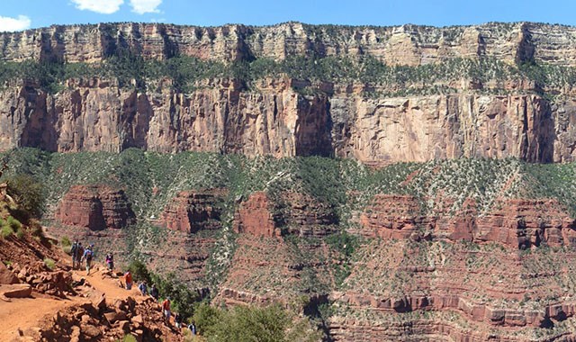 a group of hikers descending a backcountry trail with a massive wall of multi-colored cliffs in the background.