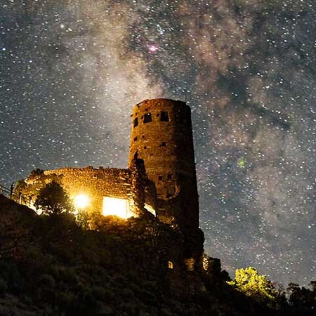 Milky Way in the sky over the Desert View Watchtower. Bright light is outlining the lower-story windows in the foreground, but the dozen or so windows in the tower on the left are dark.