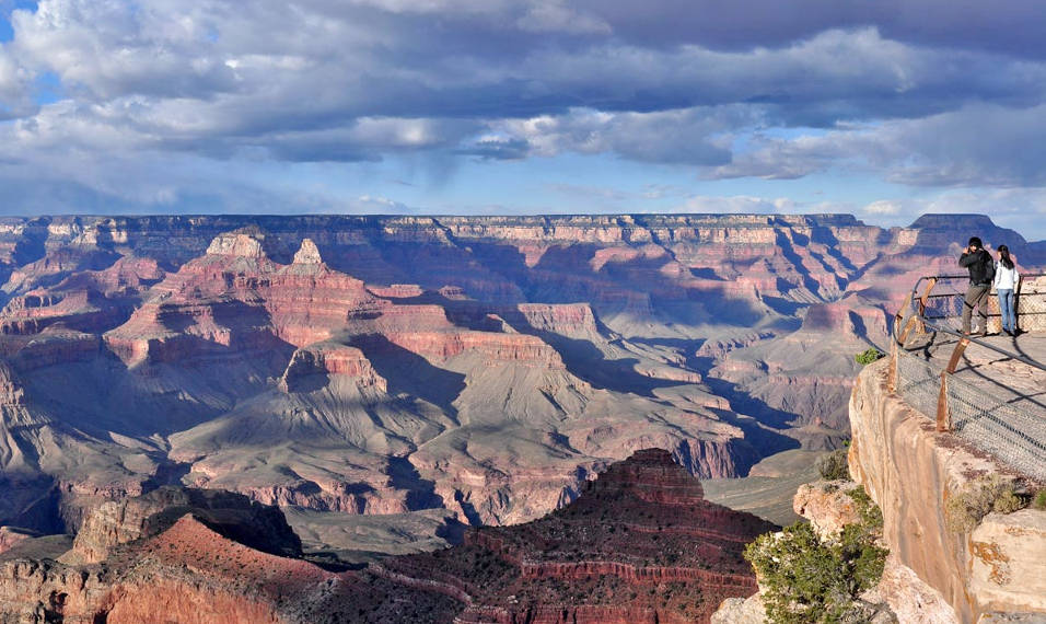 two people on the edge of a cliff and behind a metal guardrail. They are looking out at a vast and colorful landscape of stratified cliffs and peaks.