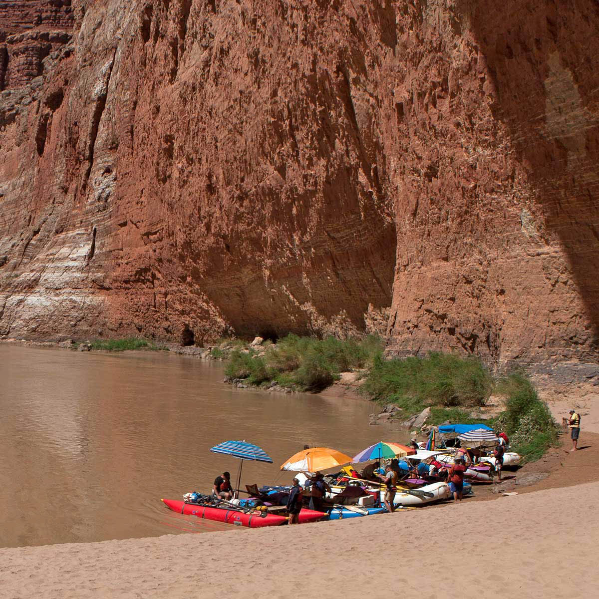 A group of rafters beneath colorful umbrellas rest at the shore next to a large cliff.