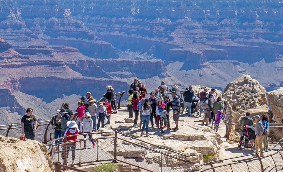 Grand Canyon National Park staff encourage all visitors to have a safe visit this holiday weekend by staying on designated trails and walkways, always keeping a safe distance from the edge of the rim, and staying behind railings and fences at overlooks. Behind metal railings at a scenic overlook, several dozen sightseers are viewing a canyon landscape