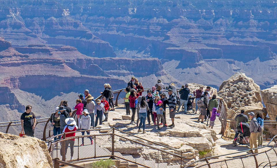 Behind metal railings at a scenic overlook, several dozen sightseers are viewing a canyon landscape