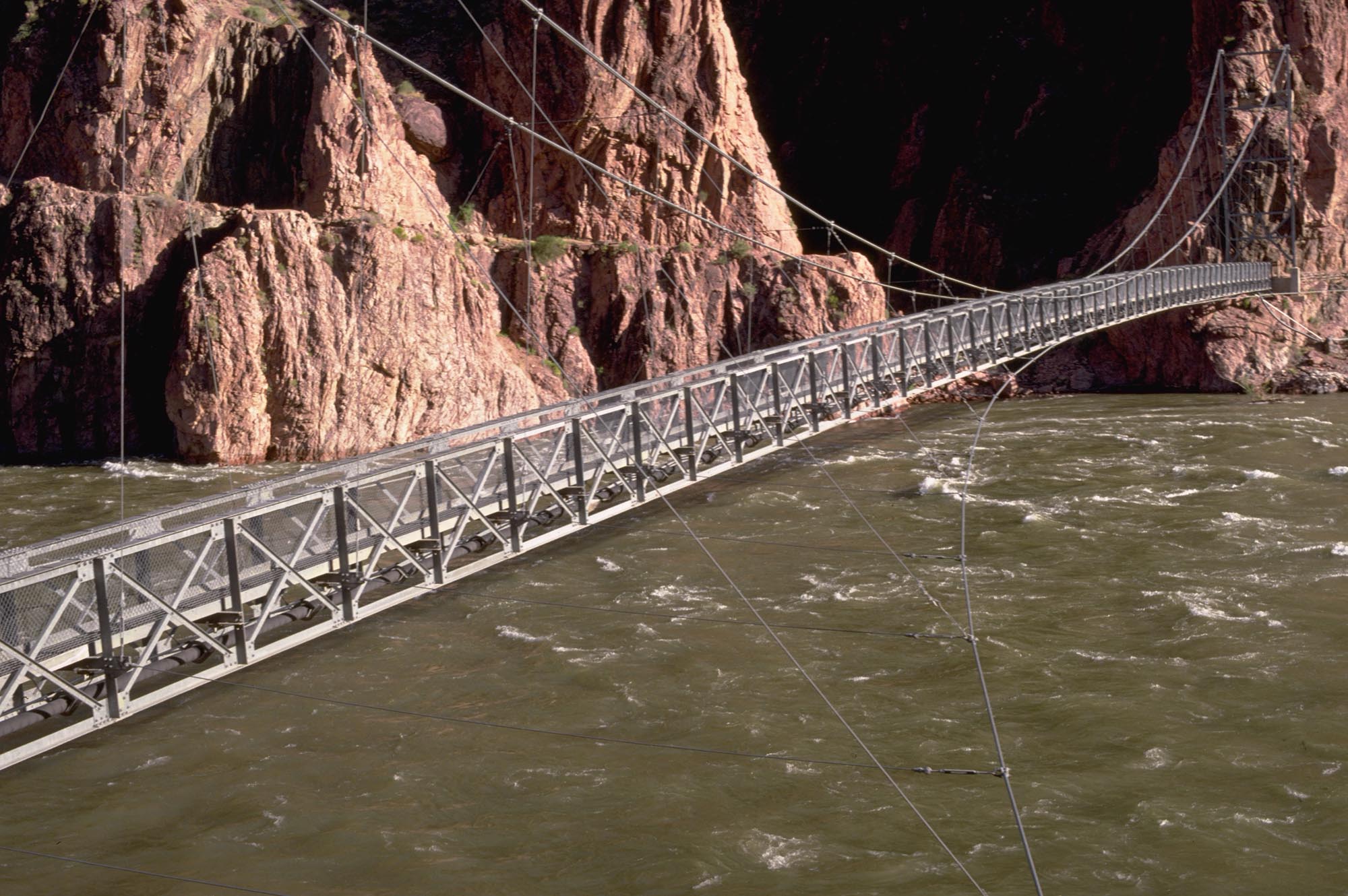 a steel suspension footbridge crossing a green river with a pipeline below the walkway.
