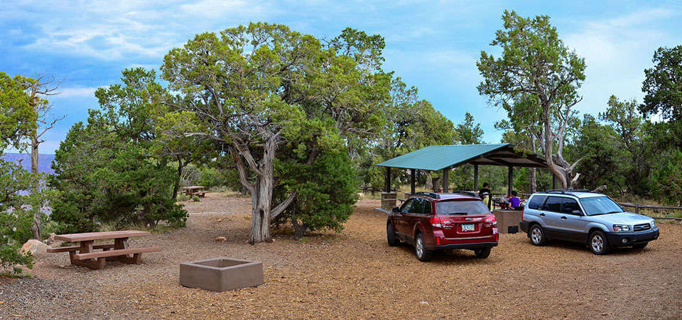 two cars parked by an open-air picnic shelter surrounded by trees. Several picnic tables and some campfire pits are visible.