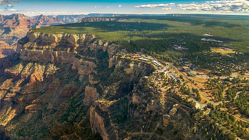 An aerial view of a village within a forested area that is on the edge of a large canyon with sheer walls.