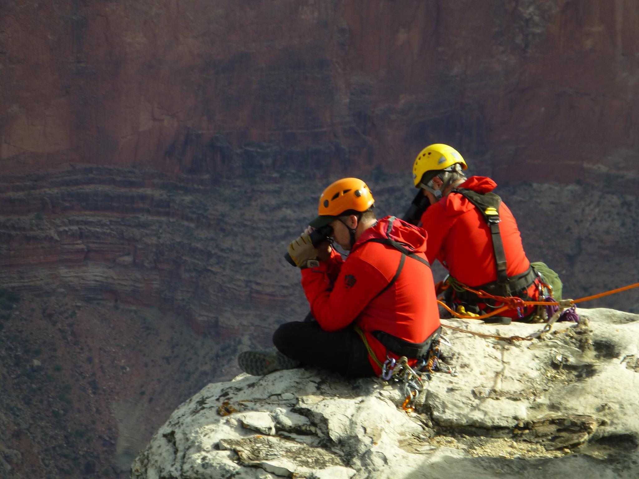 Two rangers sit at the edge of the canyon and look below the rim using binoculars