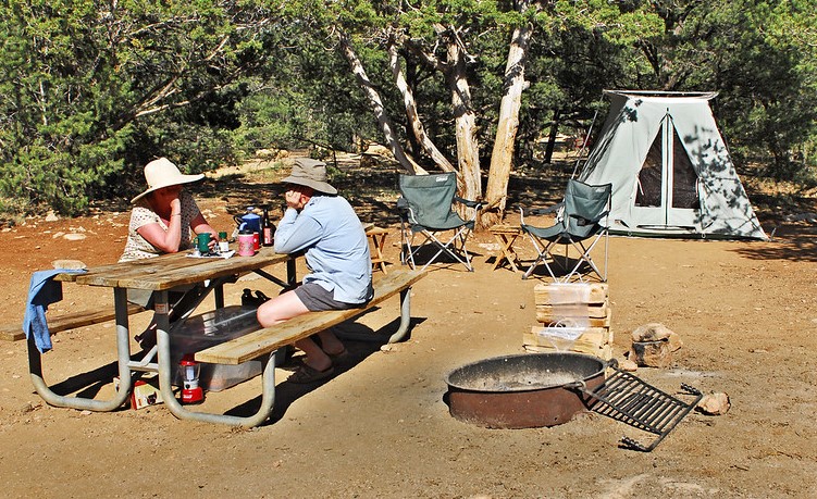 Two campers sit at a picnic table next to a fire ring
