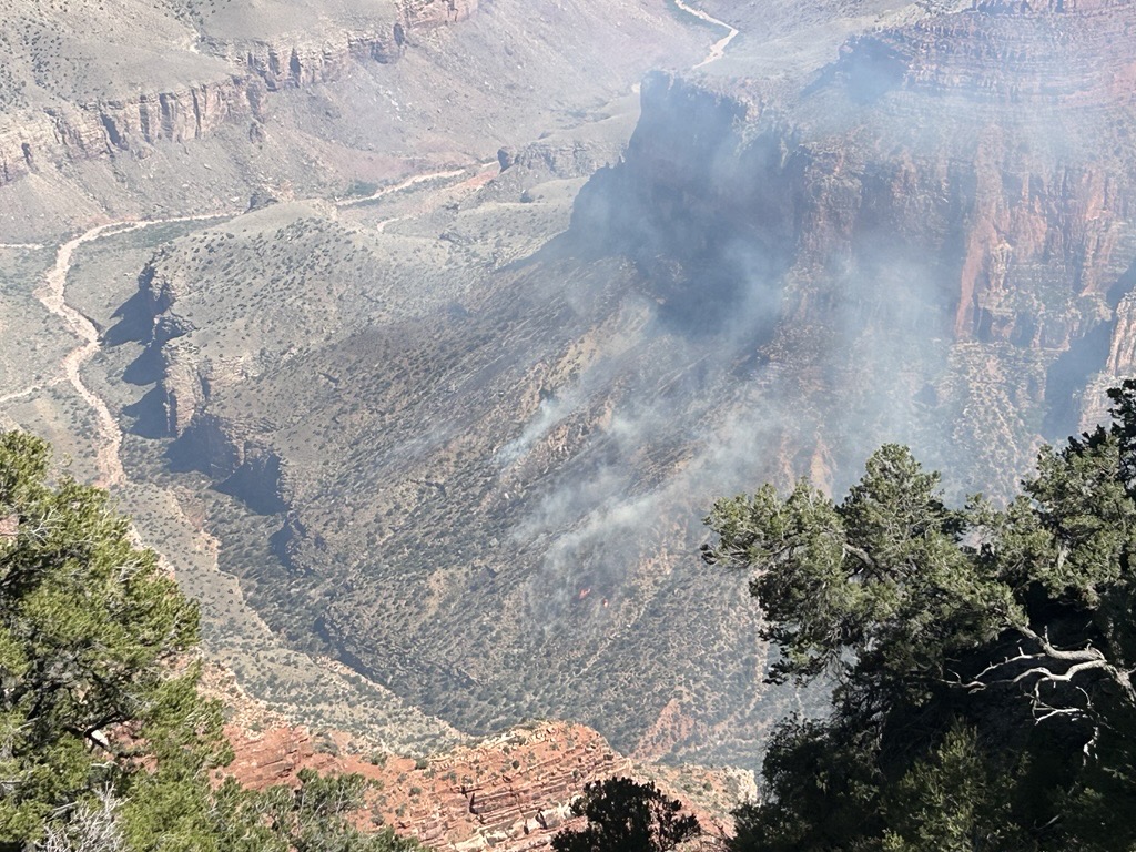 Smoke plumes drift up from a shrub-covered slope below steep cliffs.