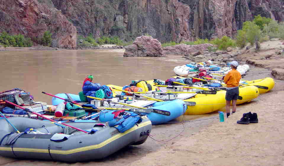 A variety of rafts representing several parties are parked on a beach by a brown river. Red and black cliffs in the background.