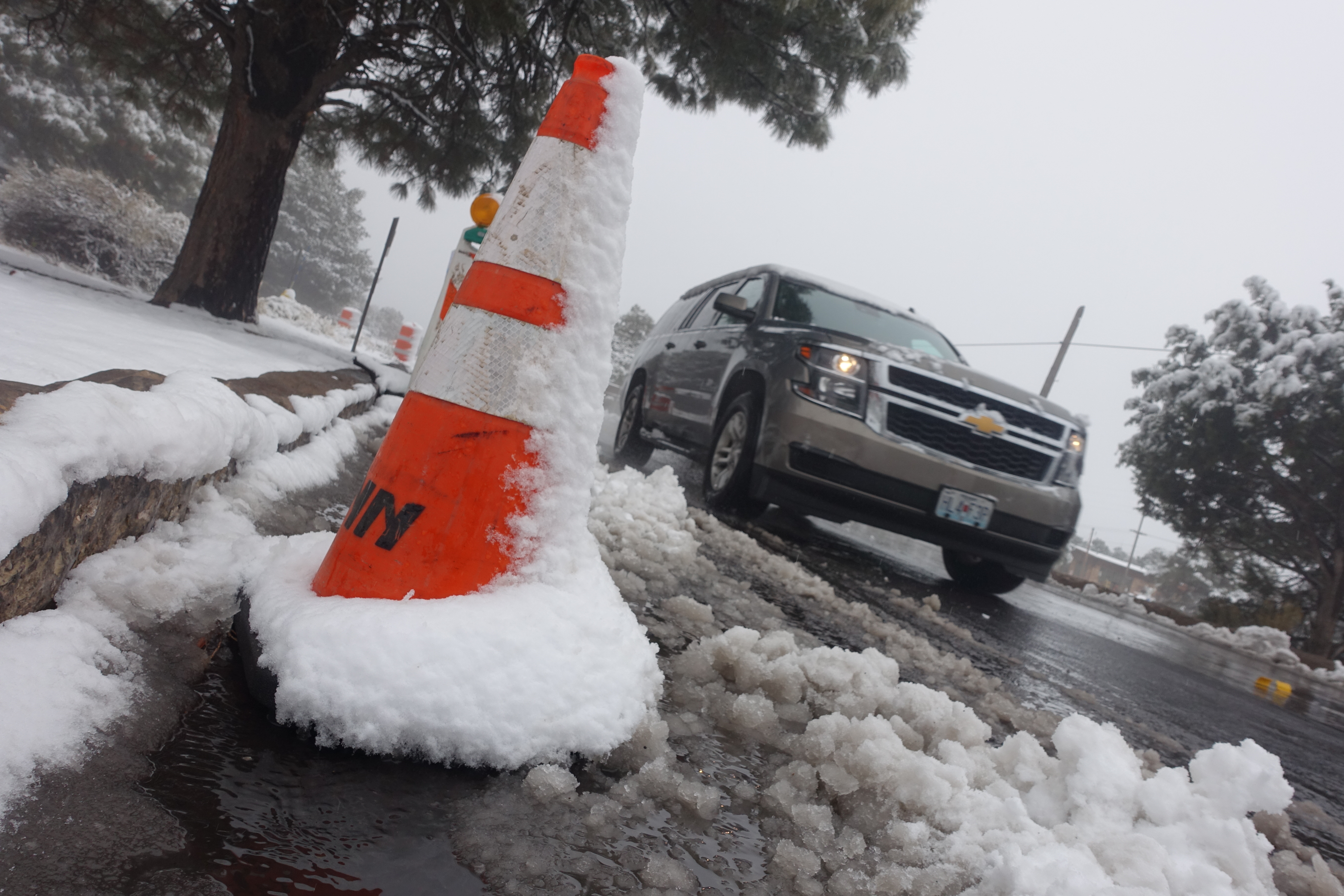 A close up of a traffic cone in the snow, in the background an SUV passes through.