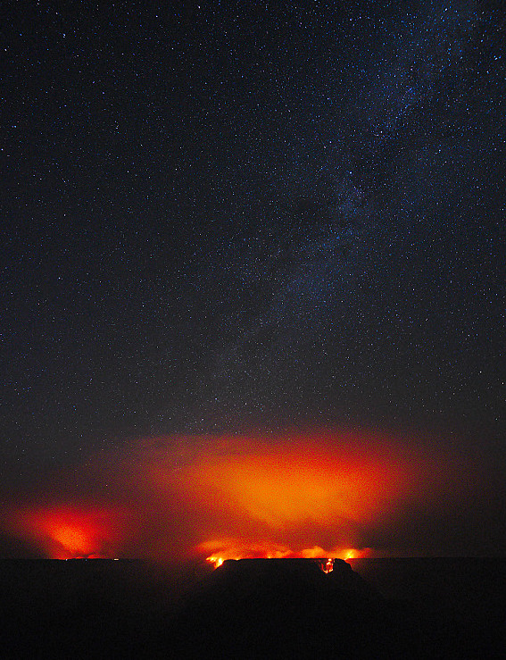 Light from wildfire flames illuminating the smoke above with an orange glow. Stars of the milky way in the dark sky above.