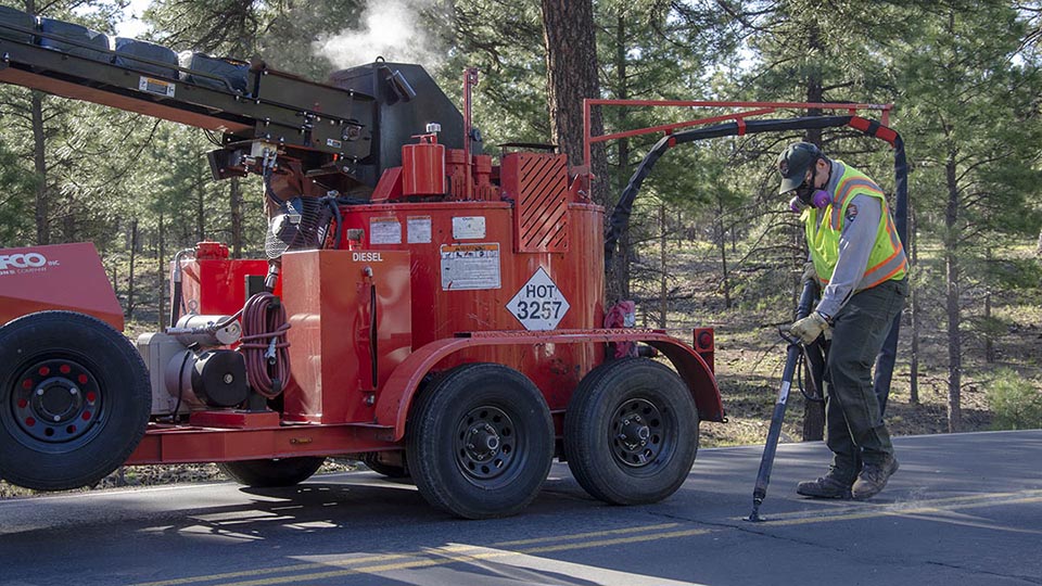 NPS road repair worker is sealing roadway cracks with an asphalt wand behind a large, red asphalt melting trailer. NPS road repair worker is sealing roadway cracks with an asphalt wand behind a large, red asphalt melting trailer.