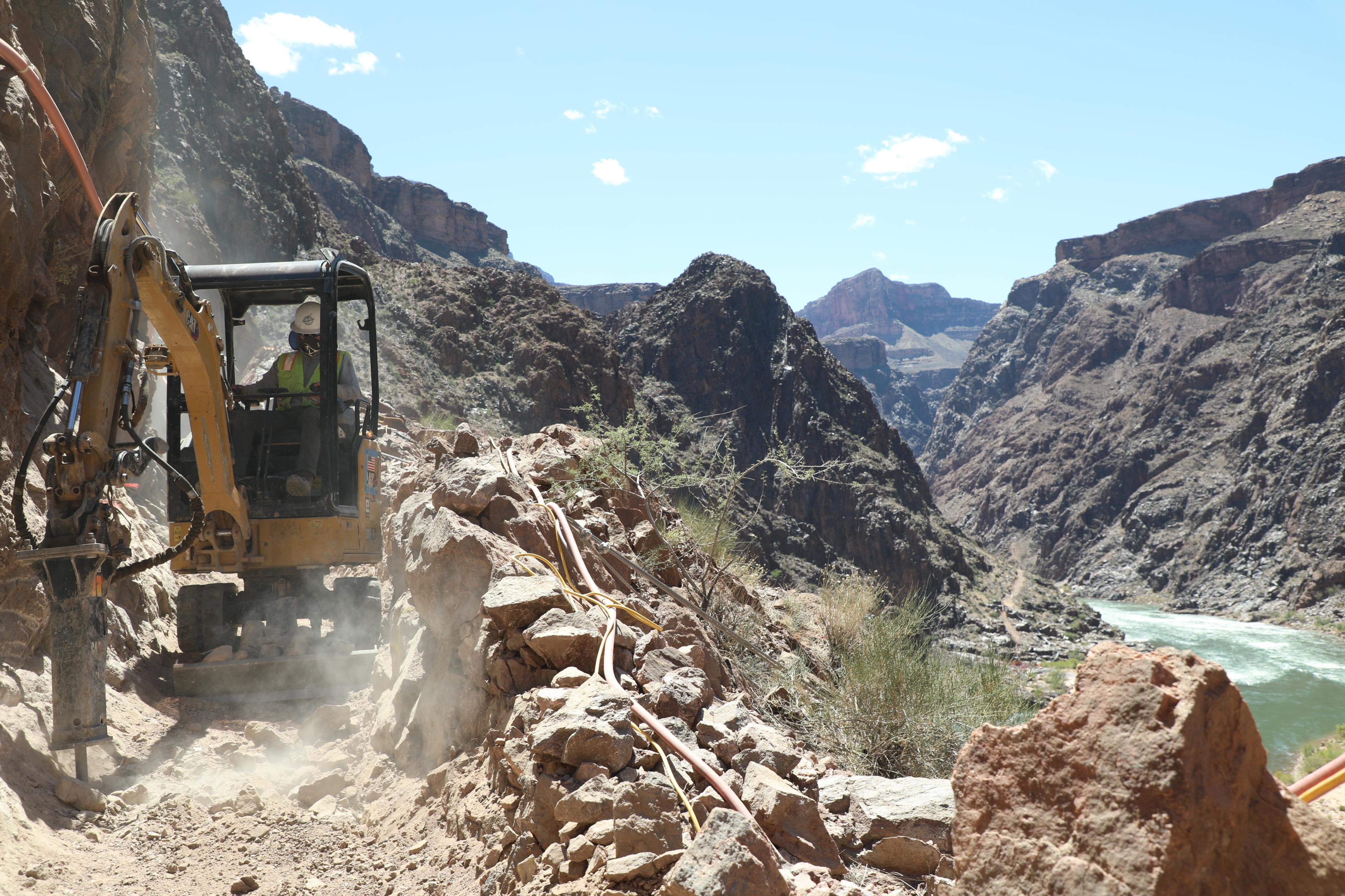 A construction bulldozer is on a trail with dust and ash. The Colorado river is in the background.