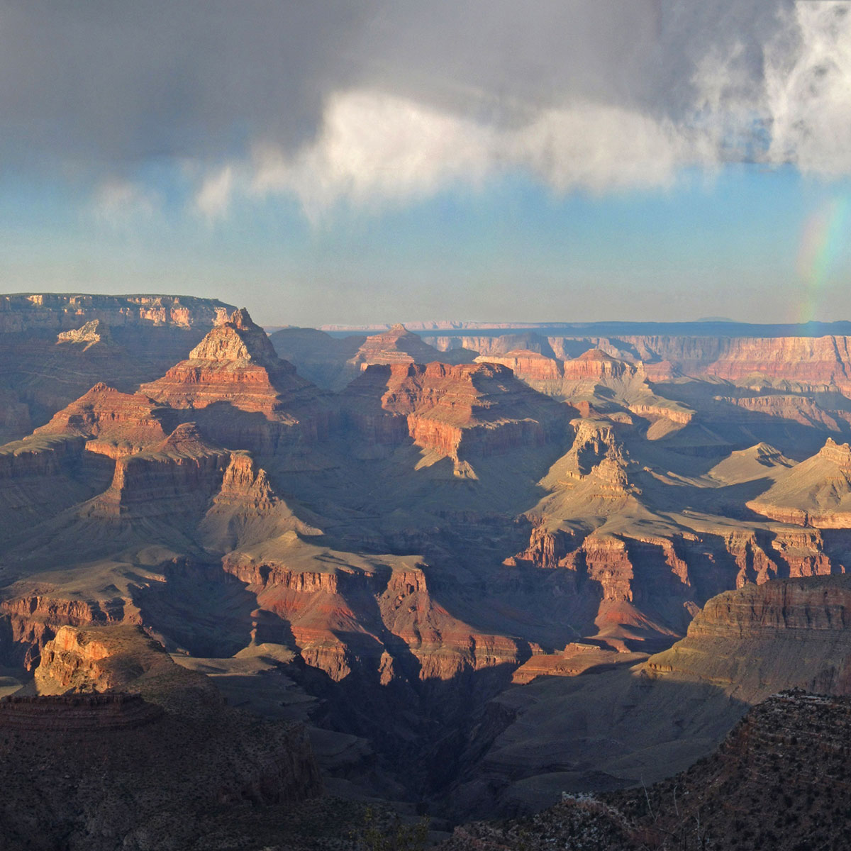 The setting sun casts shadows over the rocks with grey storm clouds above and a rainbow on the right.