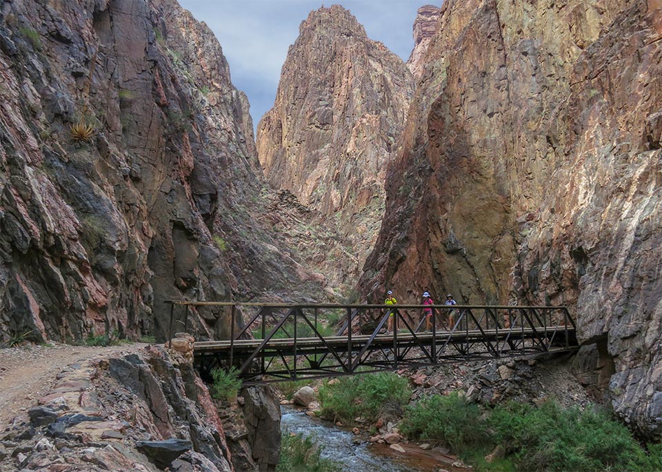 three hikers crossing a footbridge over a small creek between sheer cliff walls