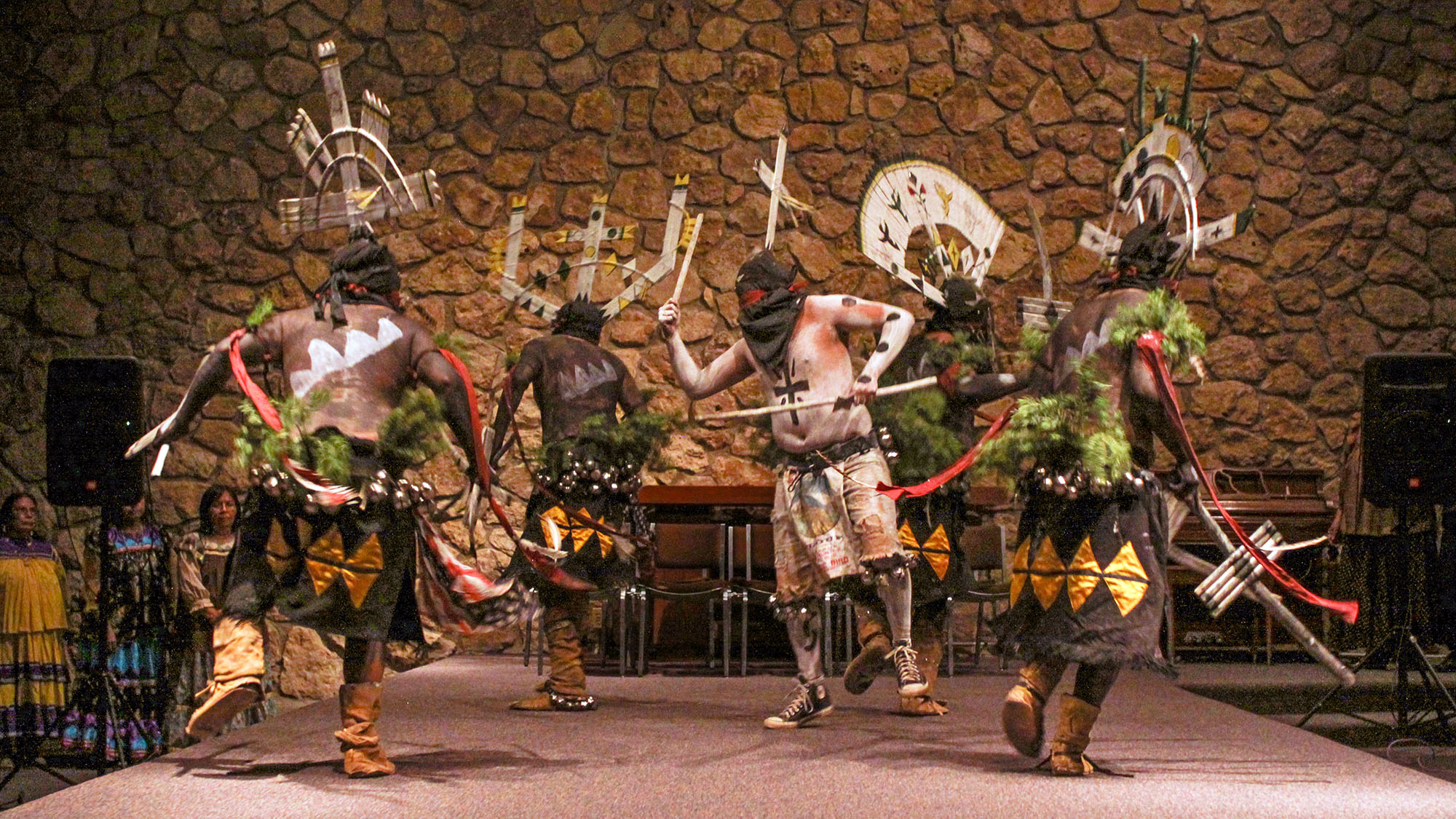 Five male Apache dancers, with designs painted on their torsos, and wearing elaborate wooden crowns, are performing on a stage in a community hall.