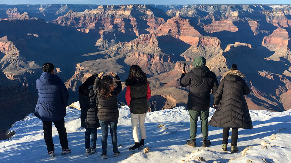 six people wearing winter clothes are standing at a snow covered scenic viewpoint overlooking a vast canyon landscape of peaks and cliffs.