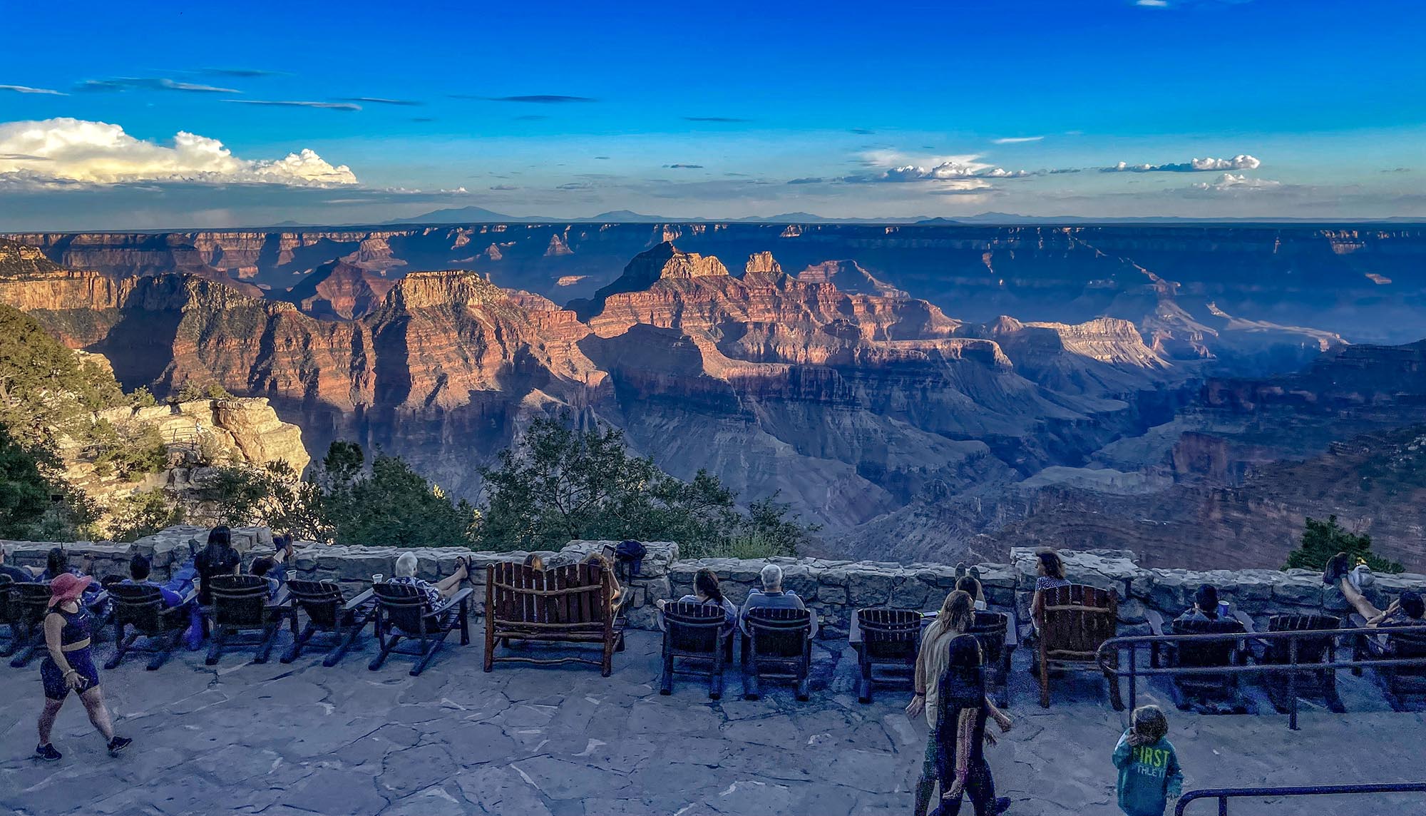 Visitors are seated on chairs on the Grand Canyon Lodge veranda, overlooking the canyon landscape at sunset