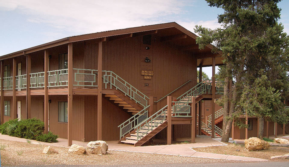 side of two-story, brown lodge building with balcony and stairways with pale green railings. Several trees taller than the building.