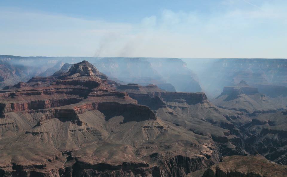 In the foreground, within an open desert landscape, a peak with stratified rock layers rises above a canyon floor. In the background, several columns of white smoke are rising, partially obscuring the view.