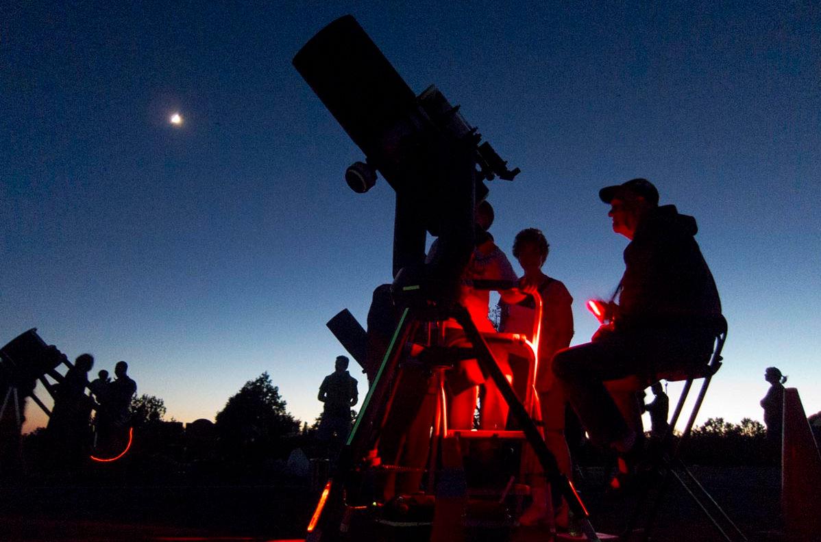 STAR PARTY - M QUINN Twilight turning into night. An astronomer is letting people look through a large telescope, that is partially illuminated by red safety lights.