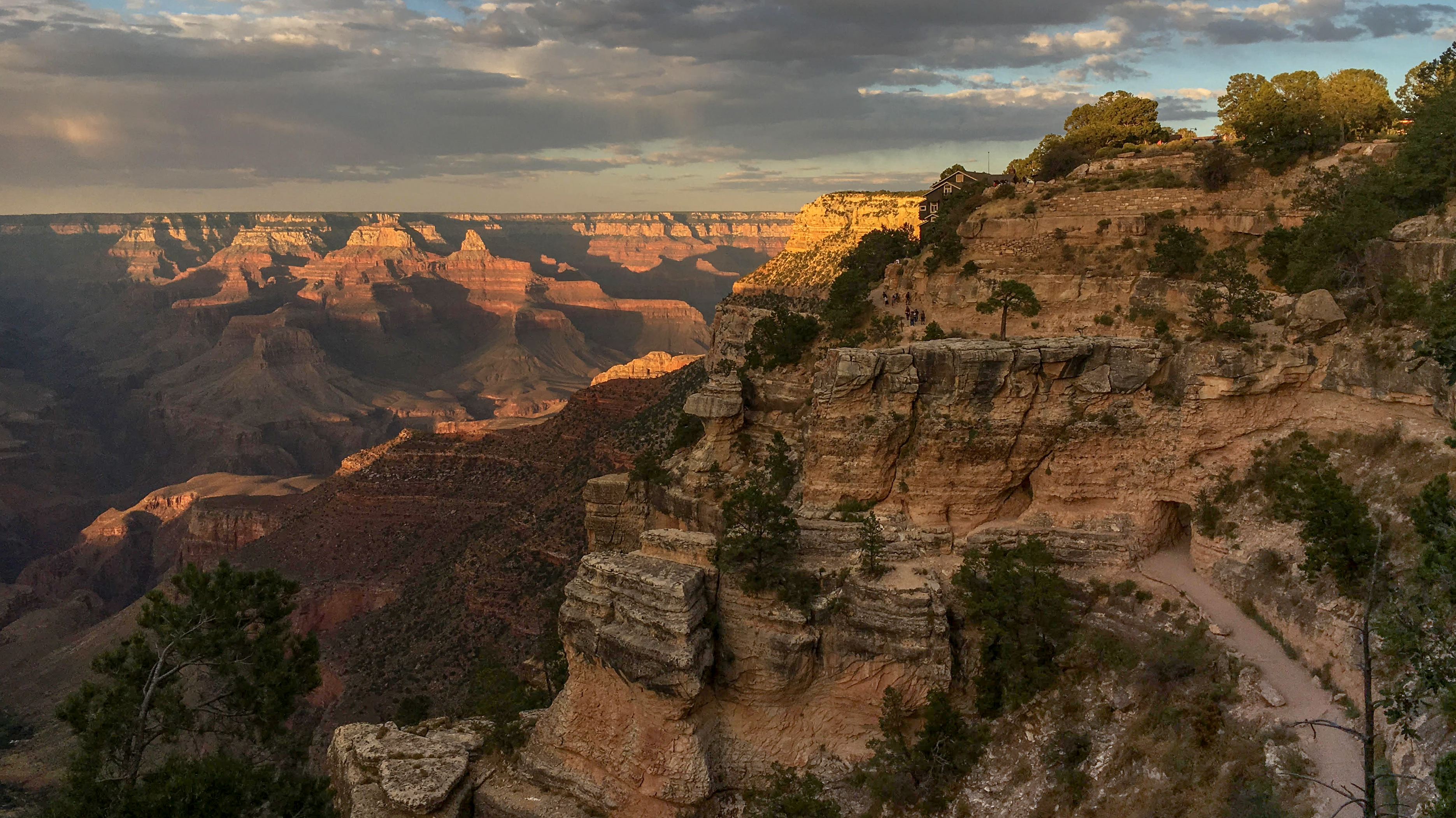 a limestone cliff in the foreground with an unpaved trail entering a tunnel in the rock. In the distance, warm sunset light falling onto peaks and cliffs within a vast canyon