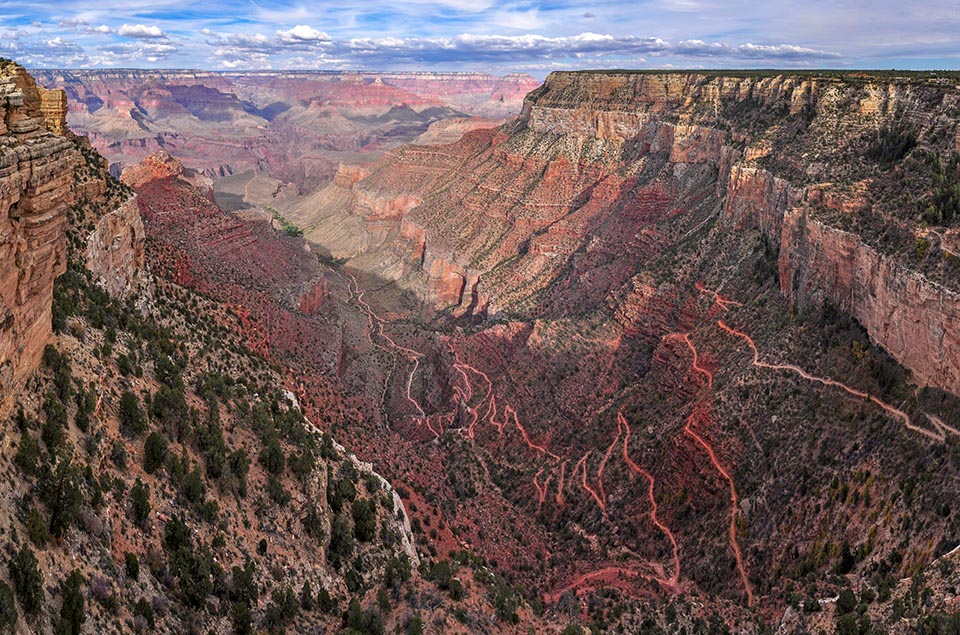 Looking down, past sheer cliffs several thousand feet, as a trail switchbacks into the depths of a colorful canyon landscape.