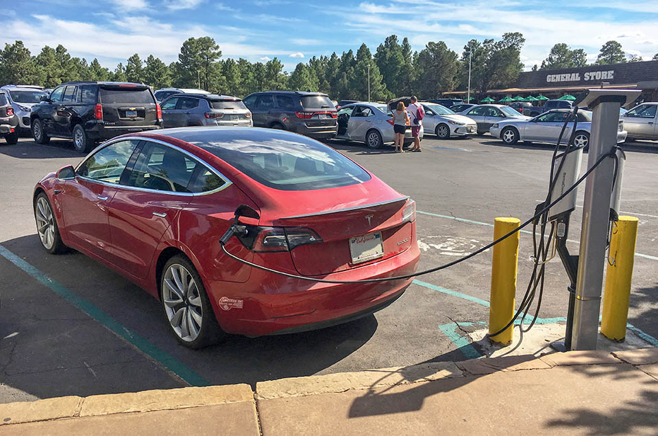 A red car plugged into a charging station at the edge of a large parking lot with many cars.