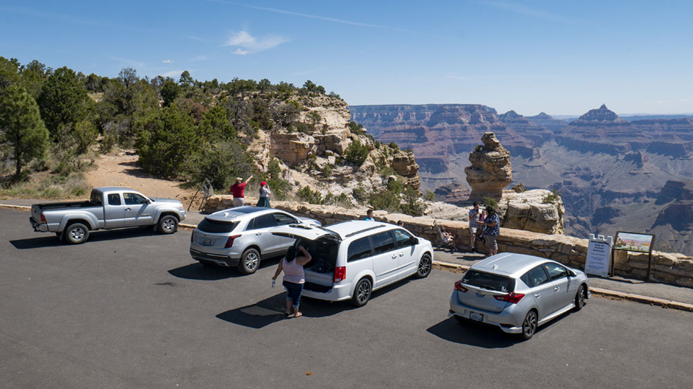 Viewing Grand Canyon from Duck on a Rock Overlook on the South Rim 4 cars parked behind a stone guard wall on the edge of a vast canyon landscape of peaks and cliffs.