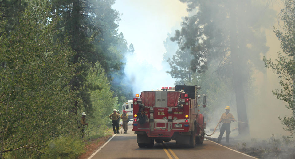 A red fire truck on a two-lane roadway. A firefighter wearing a yellow helmet and jacket is hosing down a burning snag by the side of the road. Smoke fills the air.