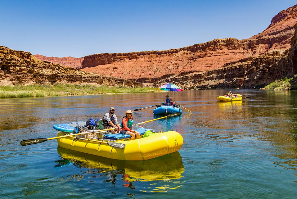 Two yellow and one blue raft floating on a stretch of calm river water, with desert cliffs in the background.