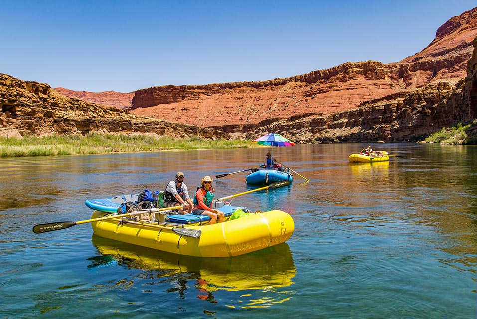 Downriver from Lees Ferry, just after starting a trip down the Colorado River through Grand Canyon National Park. Two yellow and one blue raft floating on a stretch of calm river water, with desert cliffs in the background.
