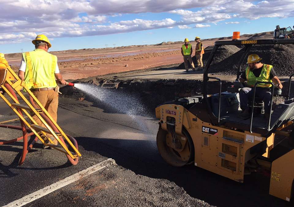 Several workers wearing yellow safety vests and helmets are cleaning up a section of paved highway that has washed away.