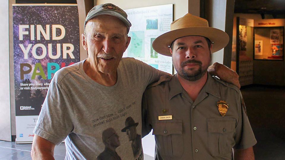 a man in his eighties standing next to a young park ranger with a short black beard at the entrance to a museum.