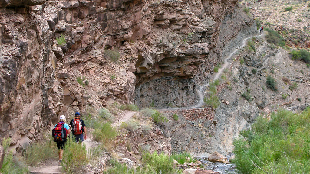 Two hikers descending a steep, unpaved, backcountry trail built into the side of a sheer cliff. On the right, a creek surrounded by green vegetation. (North Kaibab Trail) Two hikers descending a steep, unpaved, backcountry trail built into the side of a sheer cliff. On the right, a creek surrounded by green vegetation.