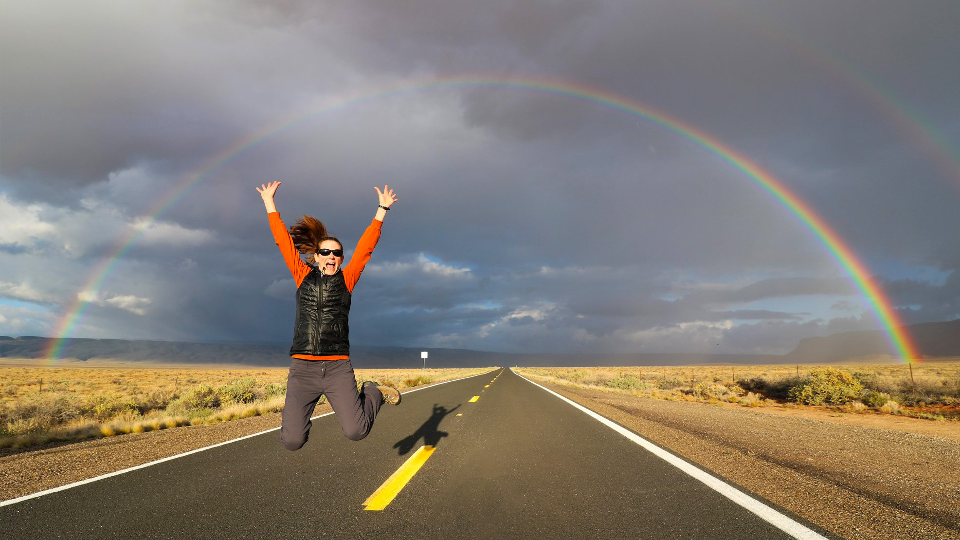 A woman is jumping for joy above a two-lane highway through a desert landscape. A full rainbow crosses the sky in a semicircular arc. A woman is jumping for joy above a two-lane highway through a desert landscape. A full rainbow crosses the sky in a semicircular arc.