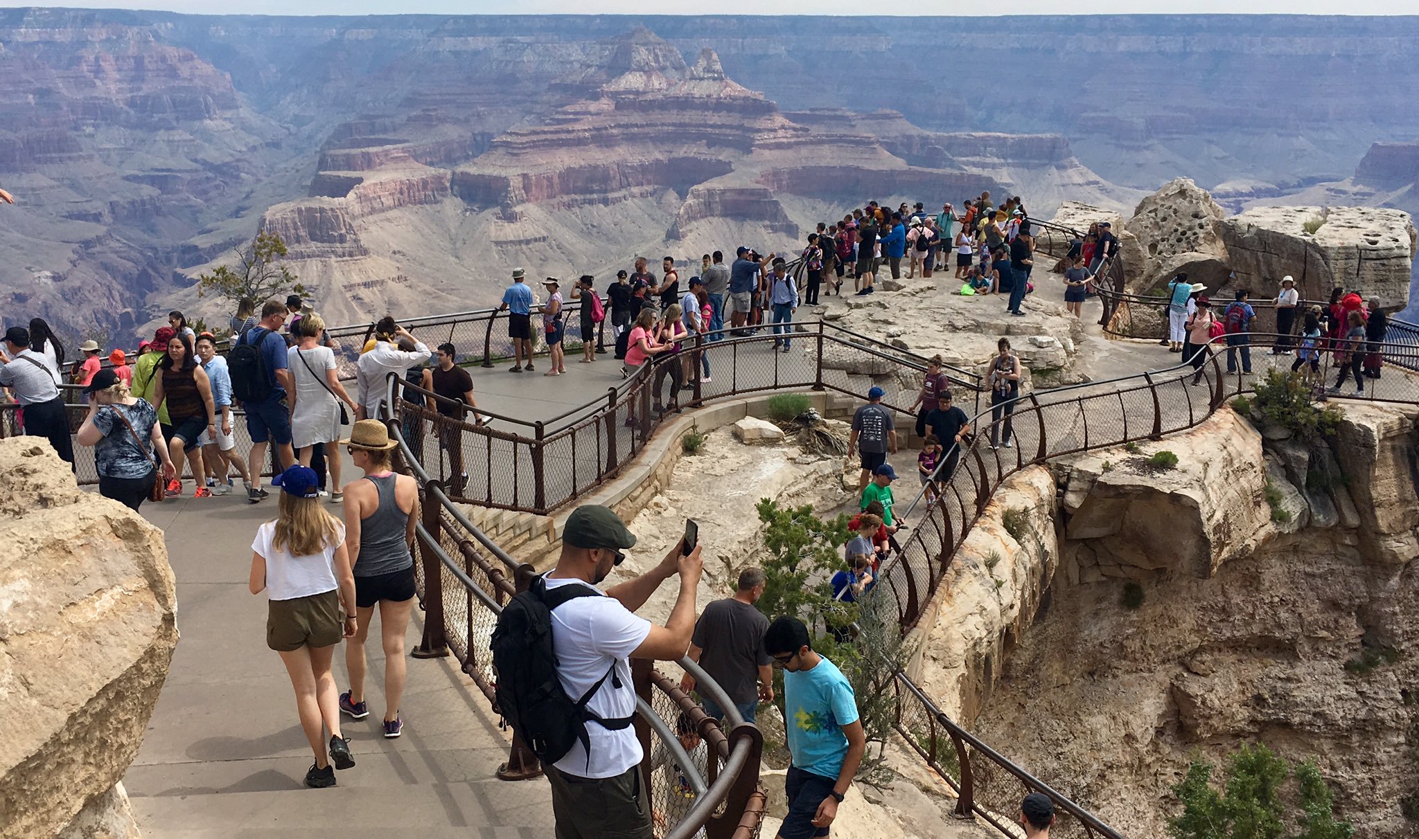 a crowd of people descending steps at a scenic overlook encircled by metal guardrails.  In the distance, an impressive peak rising out of canyon depths.