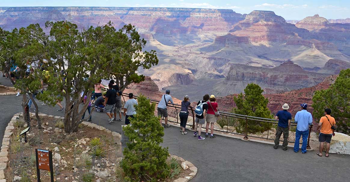 Sightseers behind a guardrail at a scenic overlook with a vast canyon landscape of cliffs and peaks in the distance.