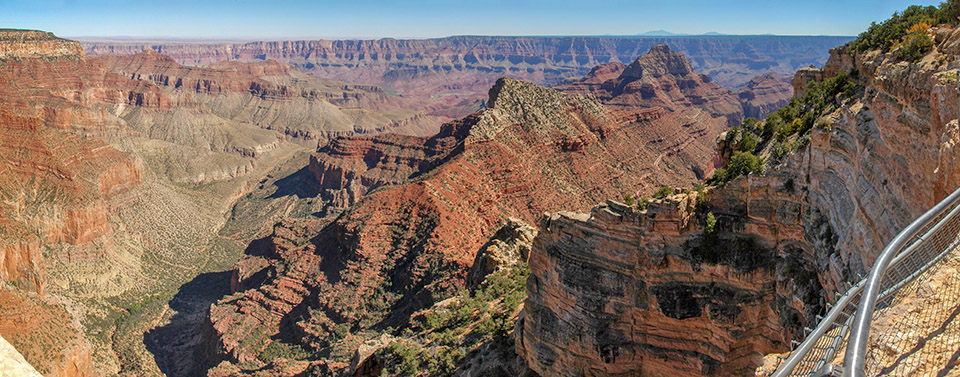 Freya Castle and Vishnu Temple as seen from the narrow walkway above Angels Window, at the eastern end of Cape Royal on the North Rim of Grand Canyon National Park. Beyond a metal guardrail, a sheer cliff gives way to colorful peaks and cliffs within a vast canyon landscape. In the distance a river is visible.
