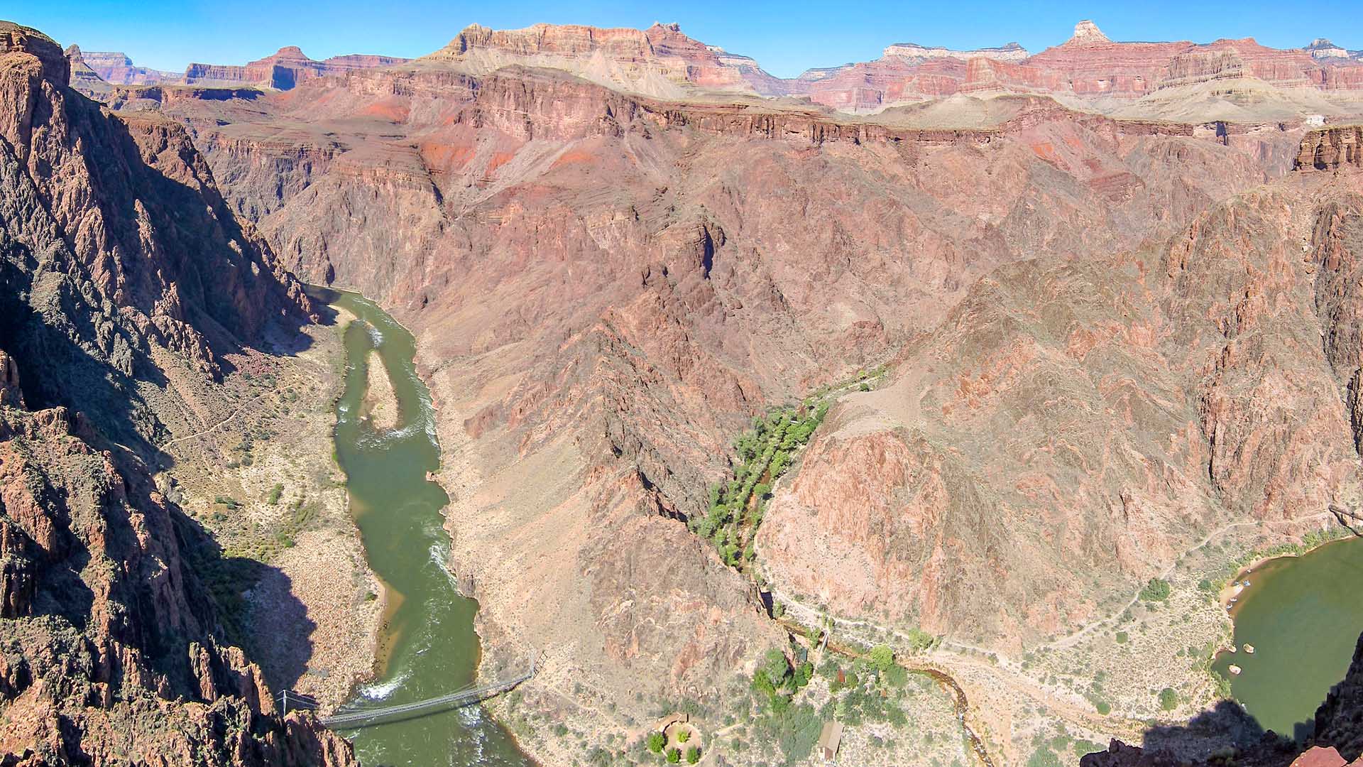Phantom Ranch Overview view down onto a green river within a rocky gorge with towering stone walls. A side canyon with trees alongside a creek