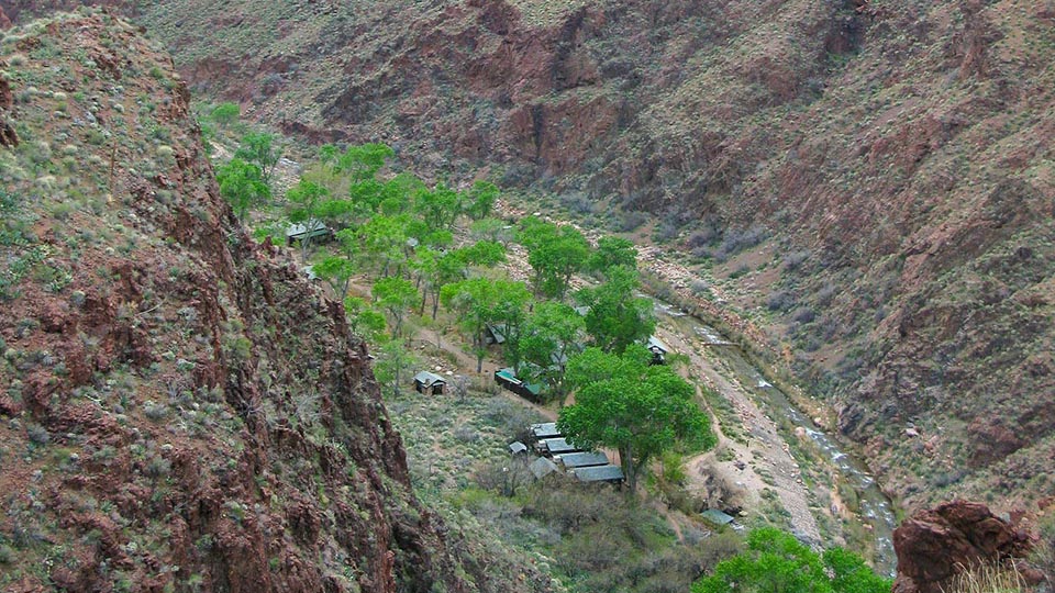 Several cabins underneath a grove of green cottonwood trees along a creek in a ravine with cliffs on both sides.