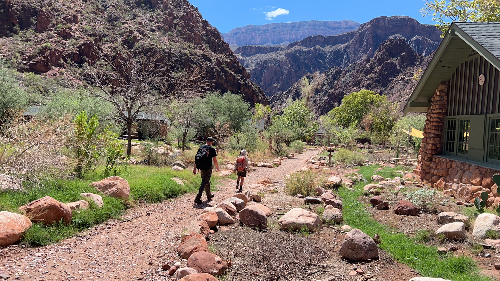 Two people walk down a trail next to a stone cabin