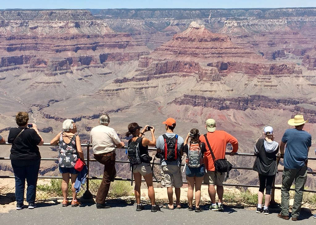 Eight Grand Canyon visitors wearing summer clothing are standing at scenic overlook behind a railing. A desert canyon landscape is in the distance.