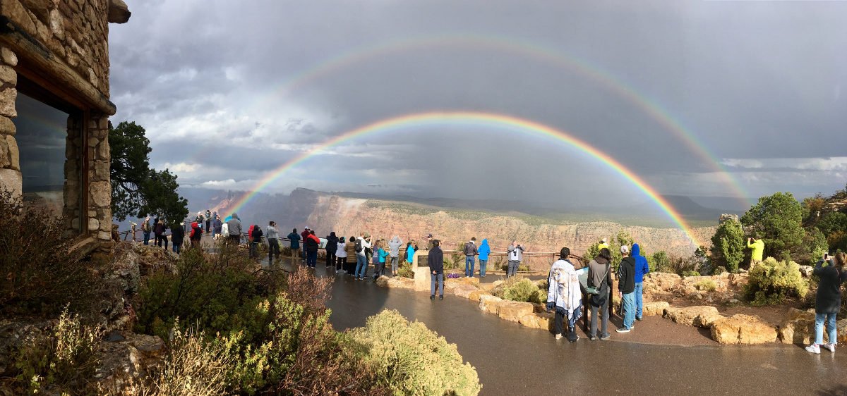 A group of sightseers standing on a wet sidewalk at a scenic overlook. In the distance a double rainbow forms a complete arc across the sky, appearing in front of a sheer cliff.