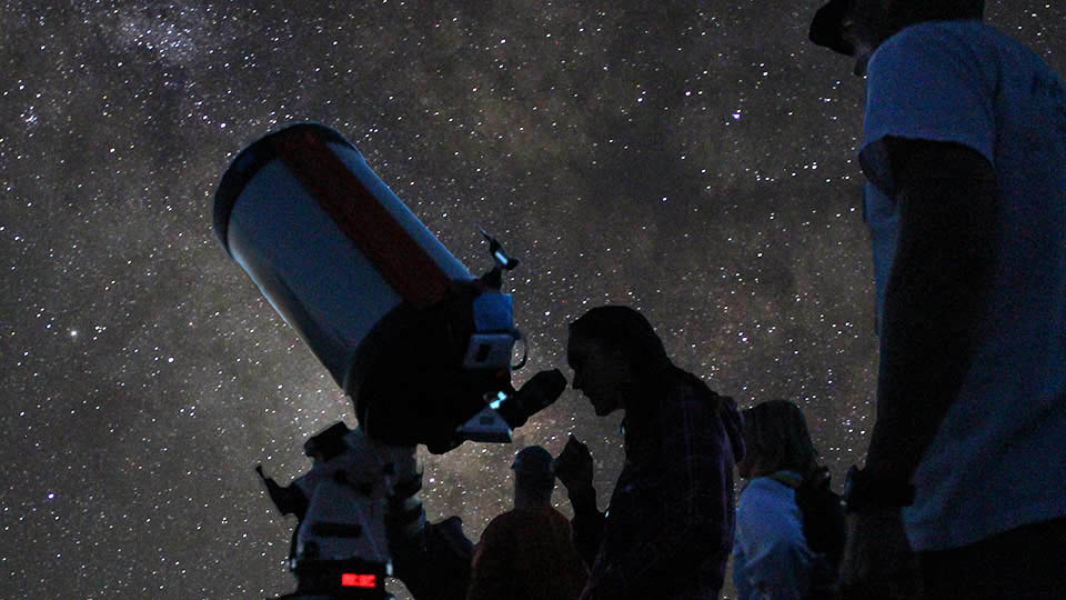 Several people looking through a large telescope in almost total darkness, with a star filled sky overhead.