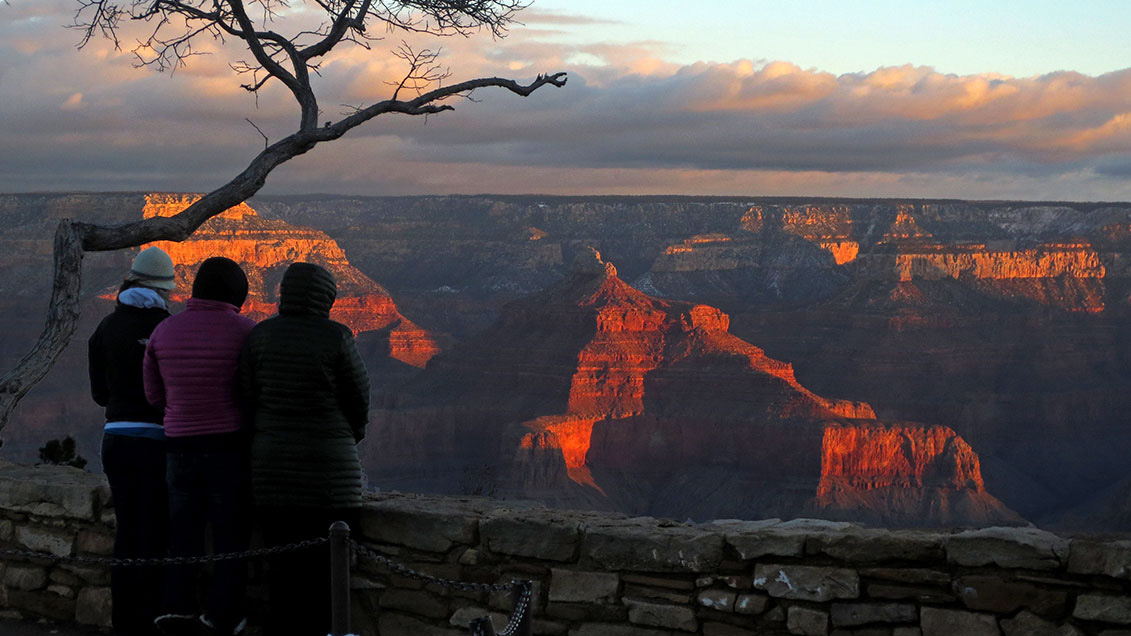 Three people wearing heavy winter coats are watching reddish sunrise light illuminating peaks and cliffs within a canyon landscape