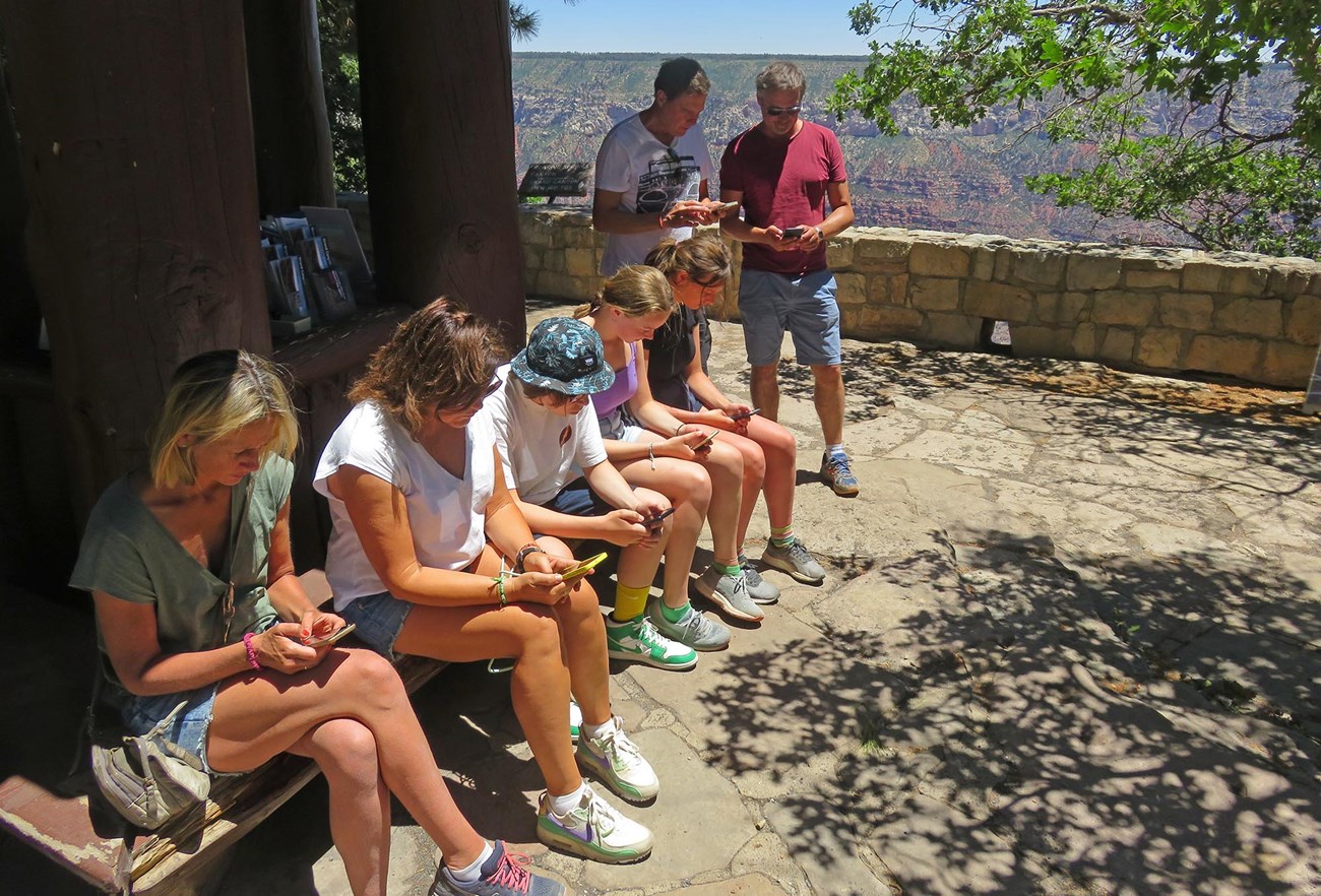 5 visitors are sitting on a bench and 2 are standing and looking at their phones beneath a shelter at a scenic overlook.