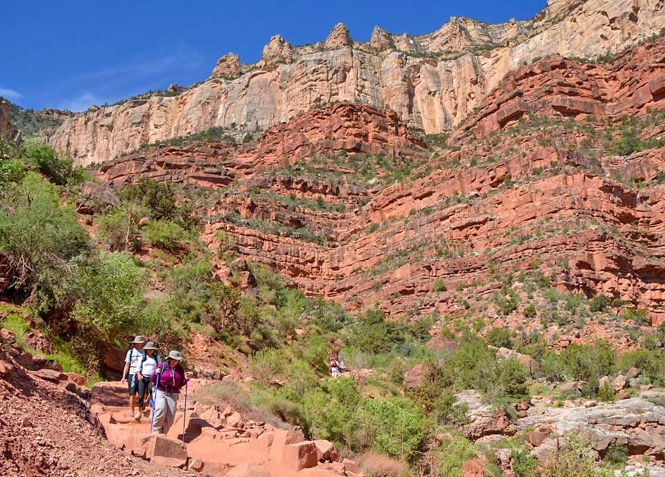 three hikers have descended several thousand feet into a canyon on a backcountry trail, and layered and colorful cliffs tower above them.